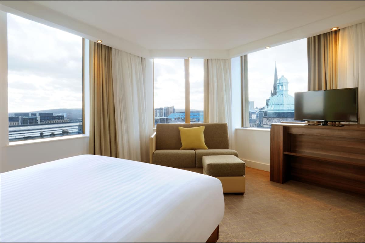 Ein Hotelzimmer mit einem großen Bett, einer Sitzecke, einem Fernseher und einem Schreibtisch. Durch große Fenster bietet sich ein Blick auf die Stadt.