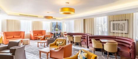 A spacious hotel lobby with a variety of seating options including sofas, armchairs, and a curved red couch. There are multiple tables and a small area with a table and chairs. The room has large windows with sheer curtains and a ceiling with recessed lighting.