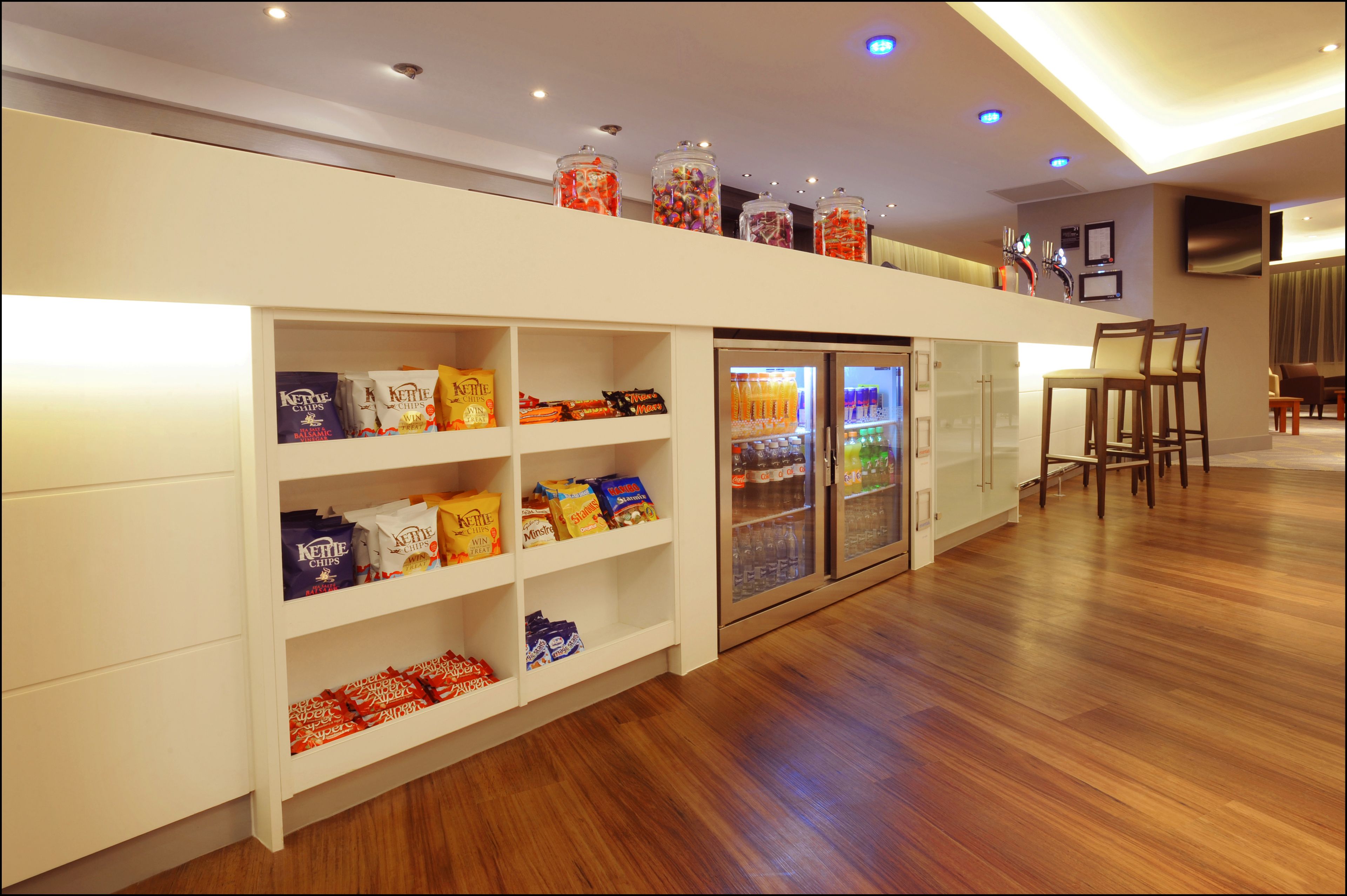 A hotel lobby with a snack bar, shelves stocked with snacks, a refrigerator with beverages, and a seating area with chairs.