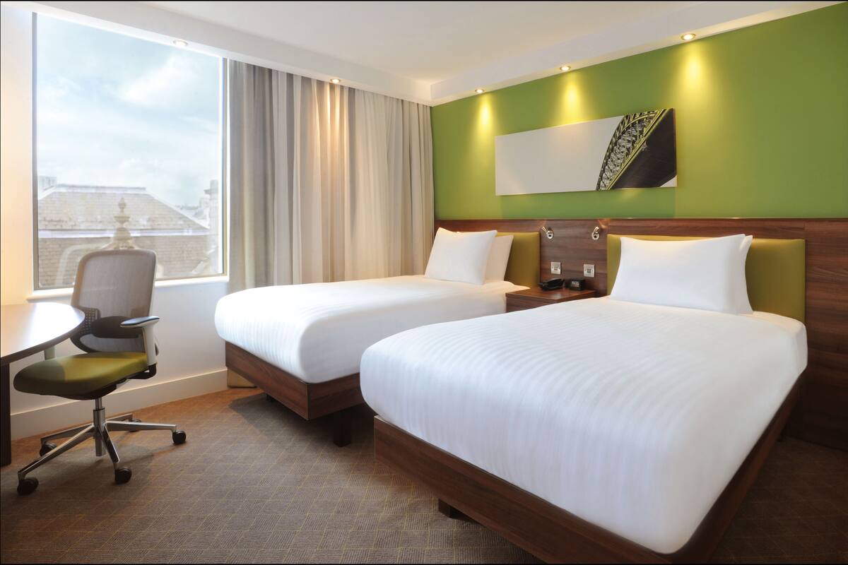 Ein Hotelzimmer mit zwei Betten, einem Schreibtisch, einem Stuhl und einem großen Fenster mit Vorhängen.