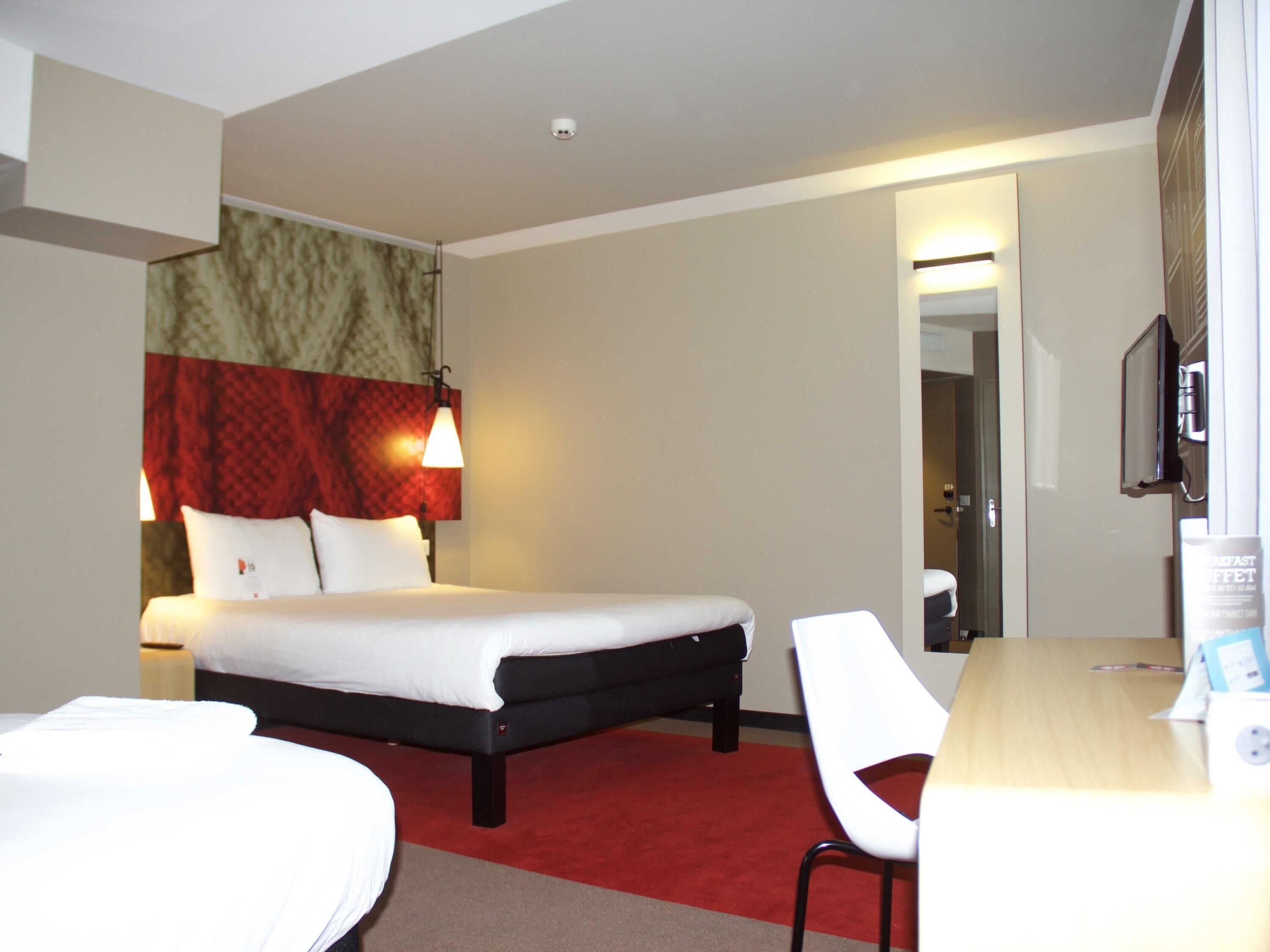 A hotel room with a bed, a desk, a chair, a TV, and a red carpet.