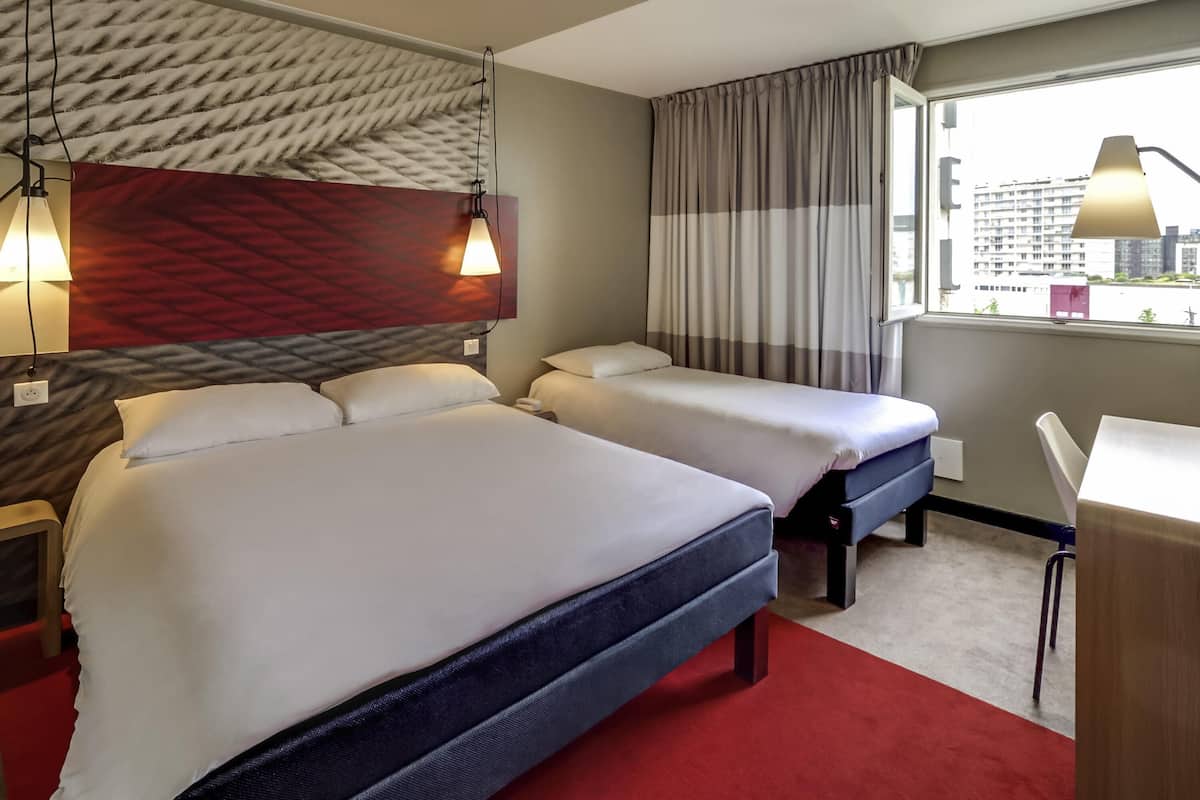 Ein Hotelzimmer mit zwei Betten, einem Schreibtisch, einem Fenster mit Vorhängen und Blick auf die Stadt.