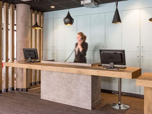 A reception desk with two HP monitors, a telephone, and a modern interior with a light-colored wall and wooden accents. - Ibis Lille Centre Grand Palais (Lille)