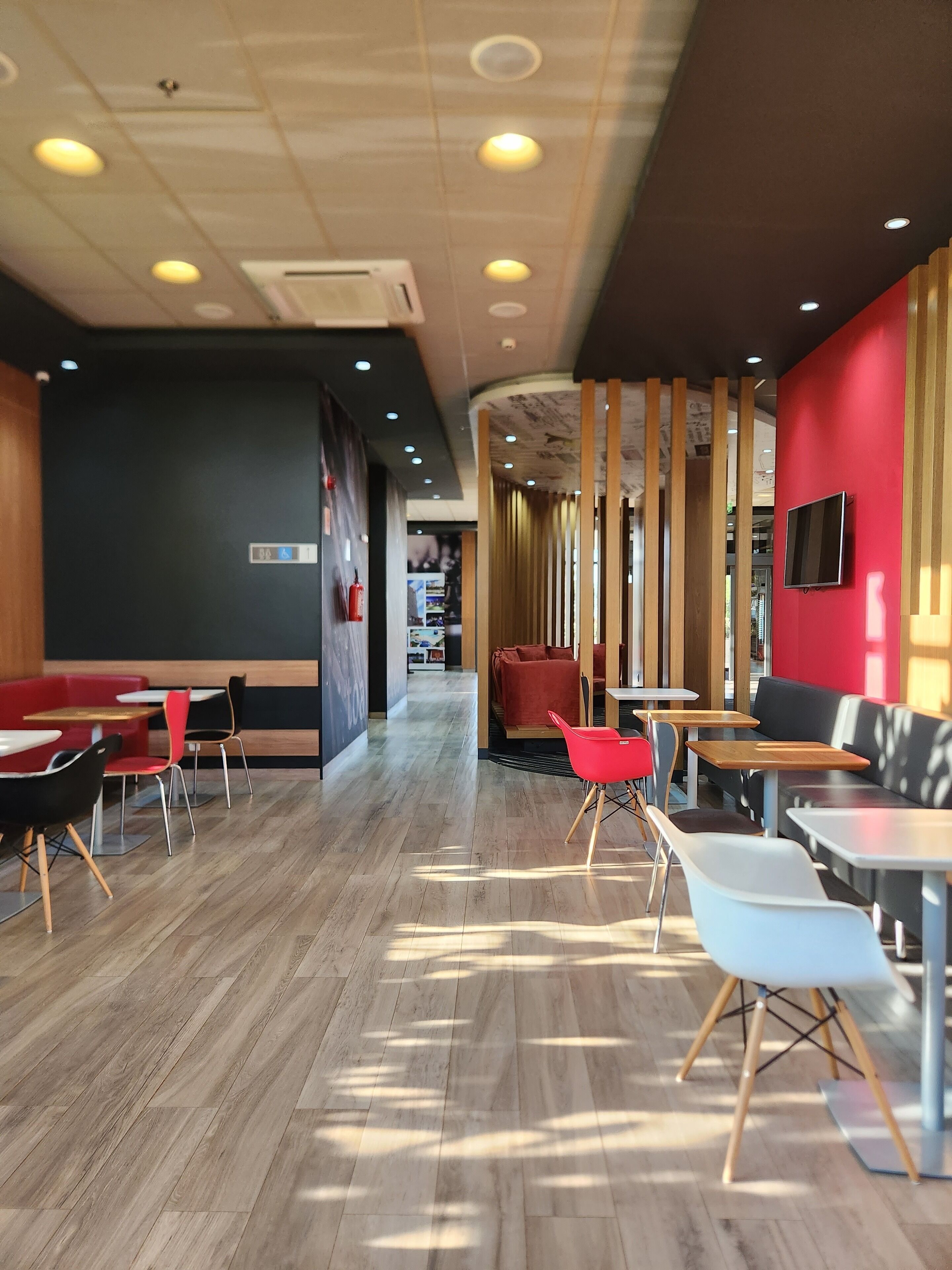 Un interior de restaurante moderno con suelo de madera, asientos en rojo y negro, y un televisor colgado en la pared.
