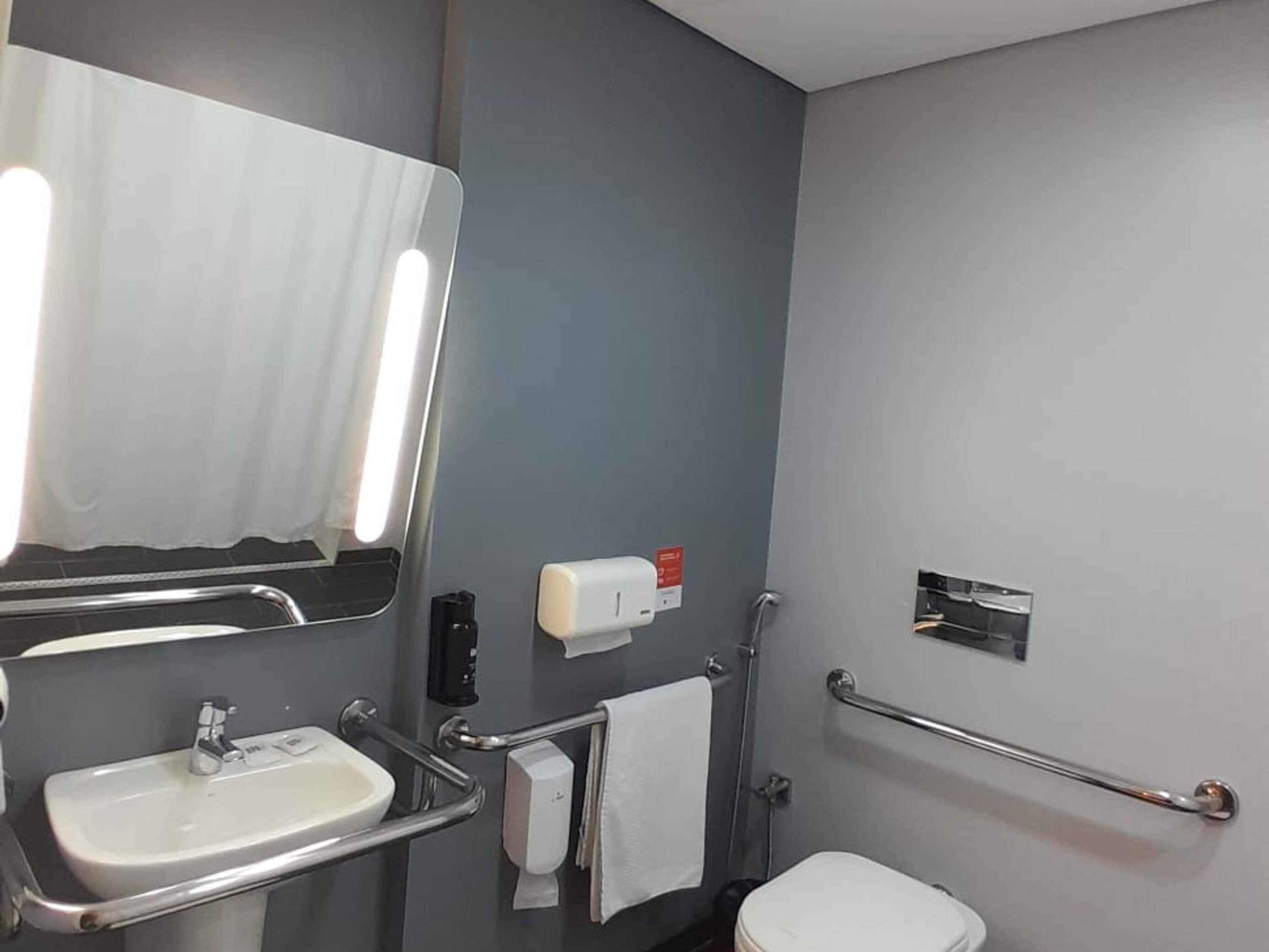 A bathroom with a sink, mirror, towel dispenser, and grab bars.