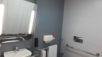 A bathroom with a sink, mirror, towel dispenser, and grab bars.