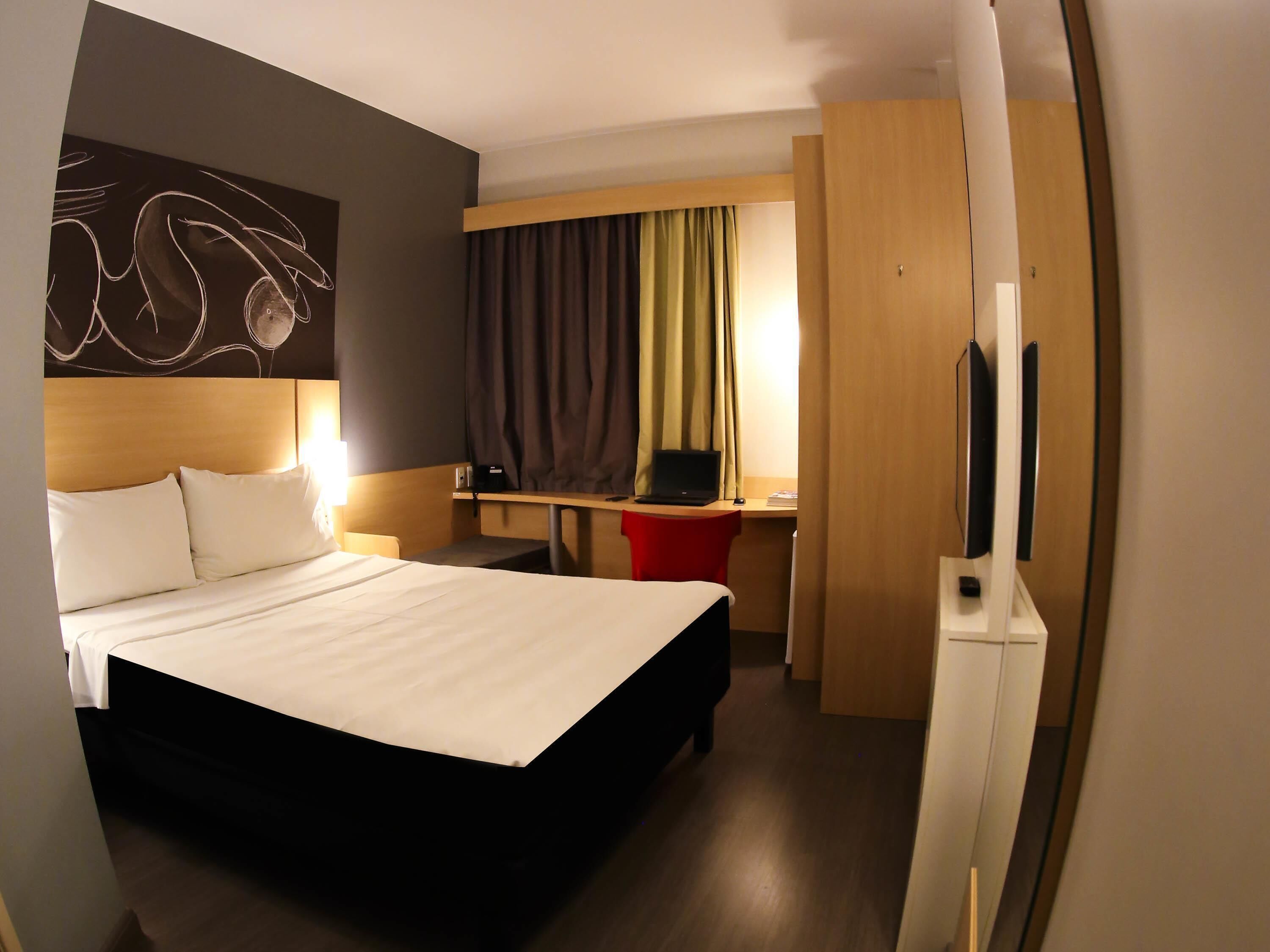 Une chambre d’hôtel avec un lit, un bureau avec un ordinateur portable, une télévision et un tableau accroché au mur.
