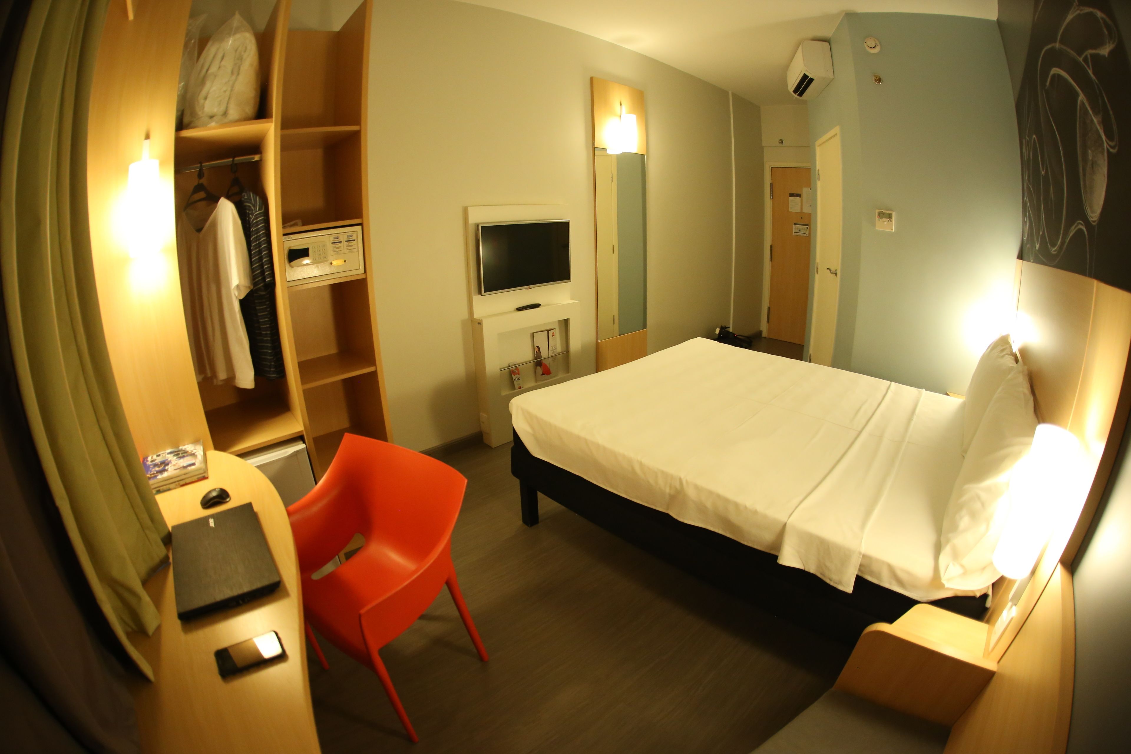 Quarto de hotel com cama, escrivaninha com computador, cadeira, televisão e guarda-roupa.