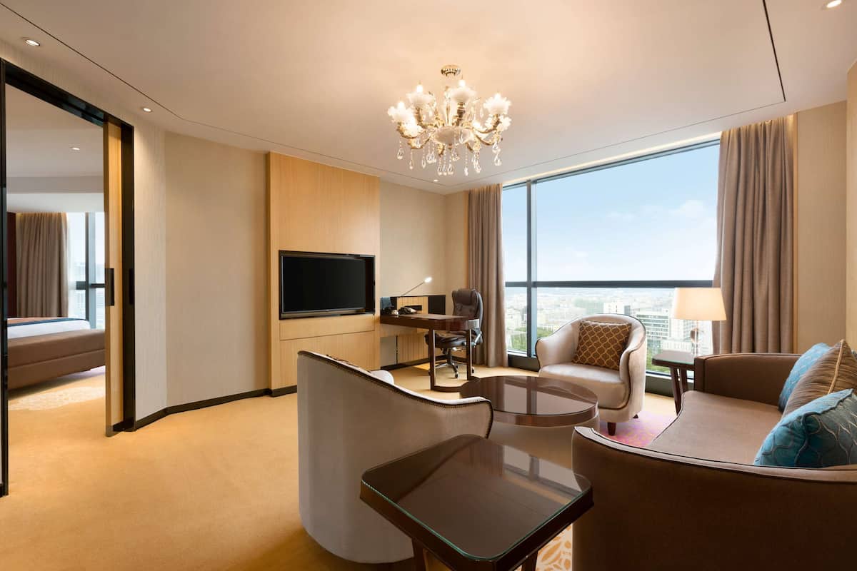 Quarto de hotel moderno com uma janela ampla, televisão de tela plana, escrivaninha com cadeira, sofá e lustre.