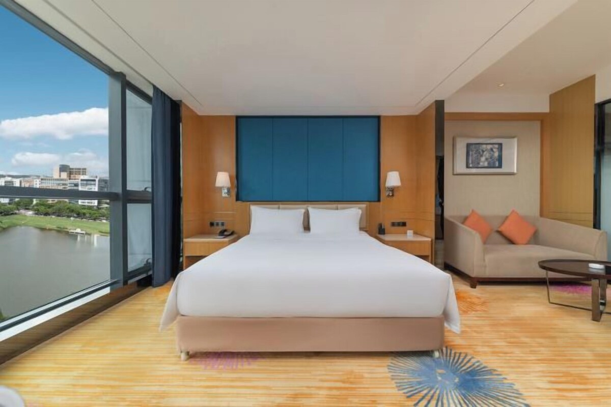 A modern hotel room with a large bed, a sofa, a small table, and a view of a body of water.