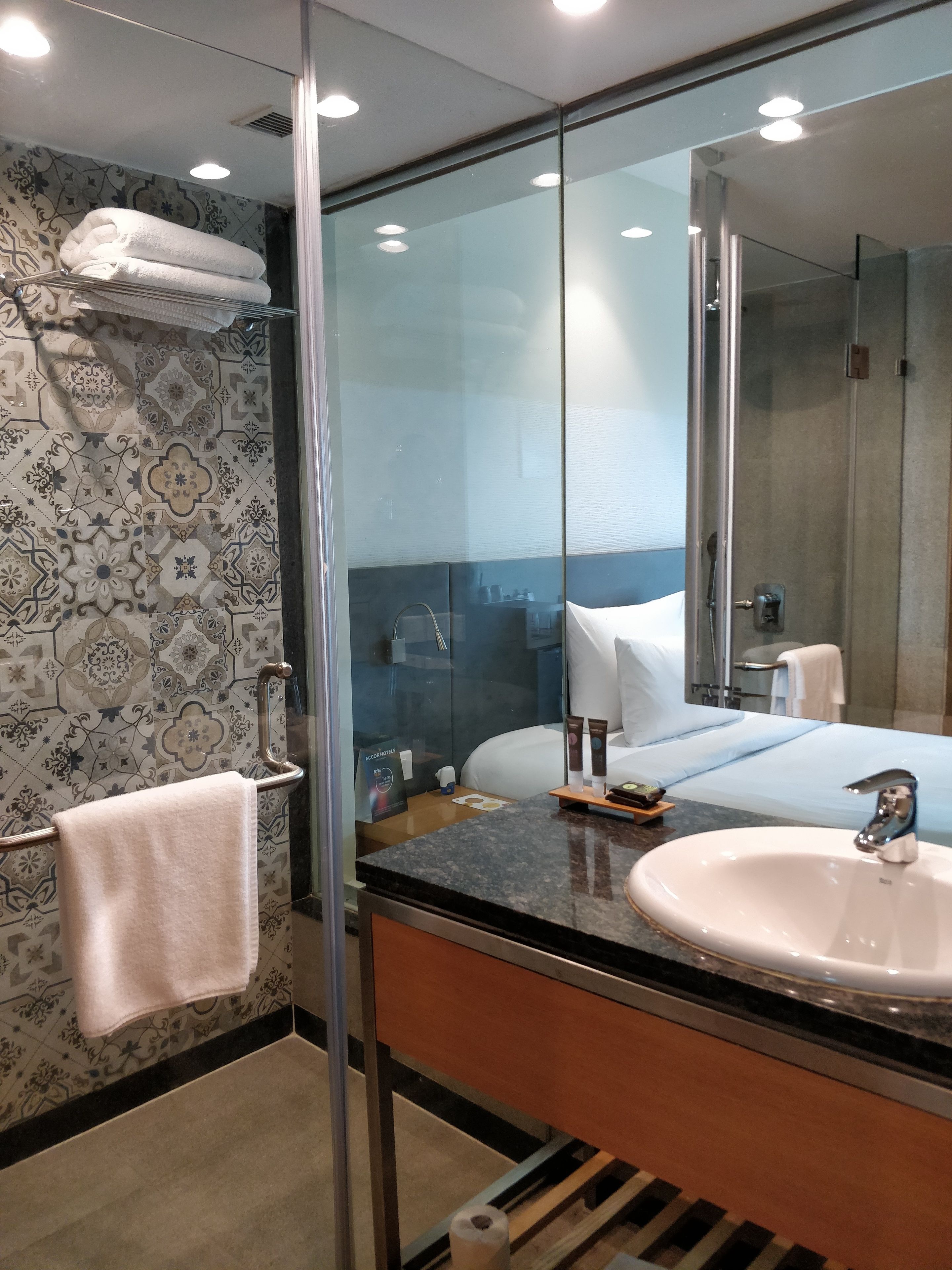 A modern bathroom with a glass shower enclosure, a large mirror, a sink, and a neatly made bed.