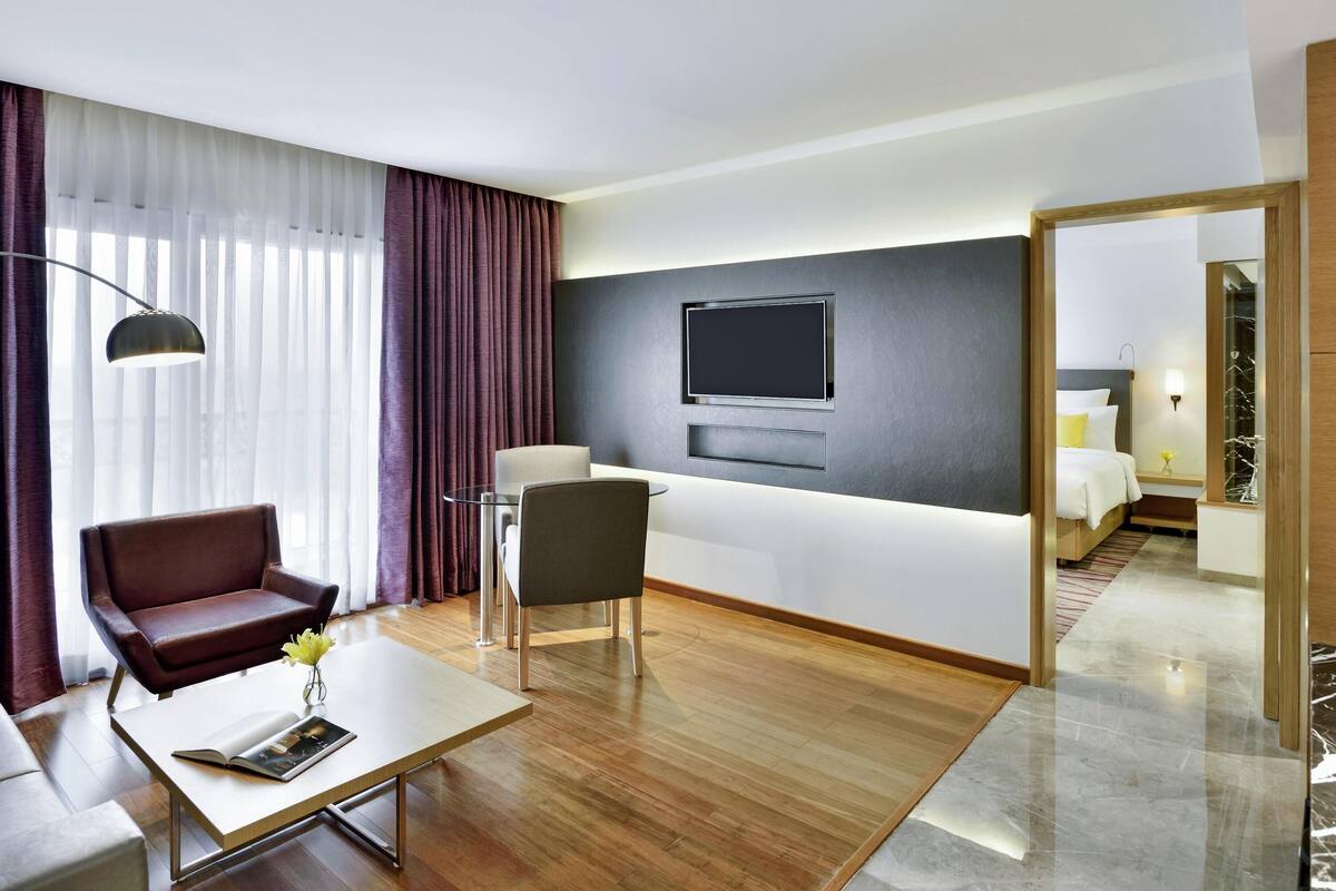 A modern hotel room with a wooden floor, a sofa, a chair, a TV mounted on the wall, and a bed with a nightstand.
