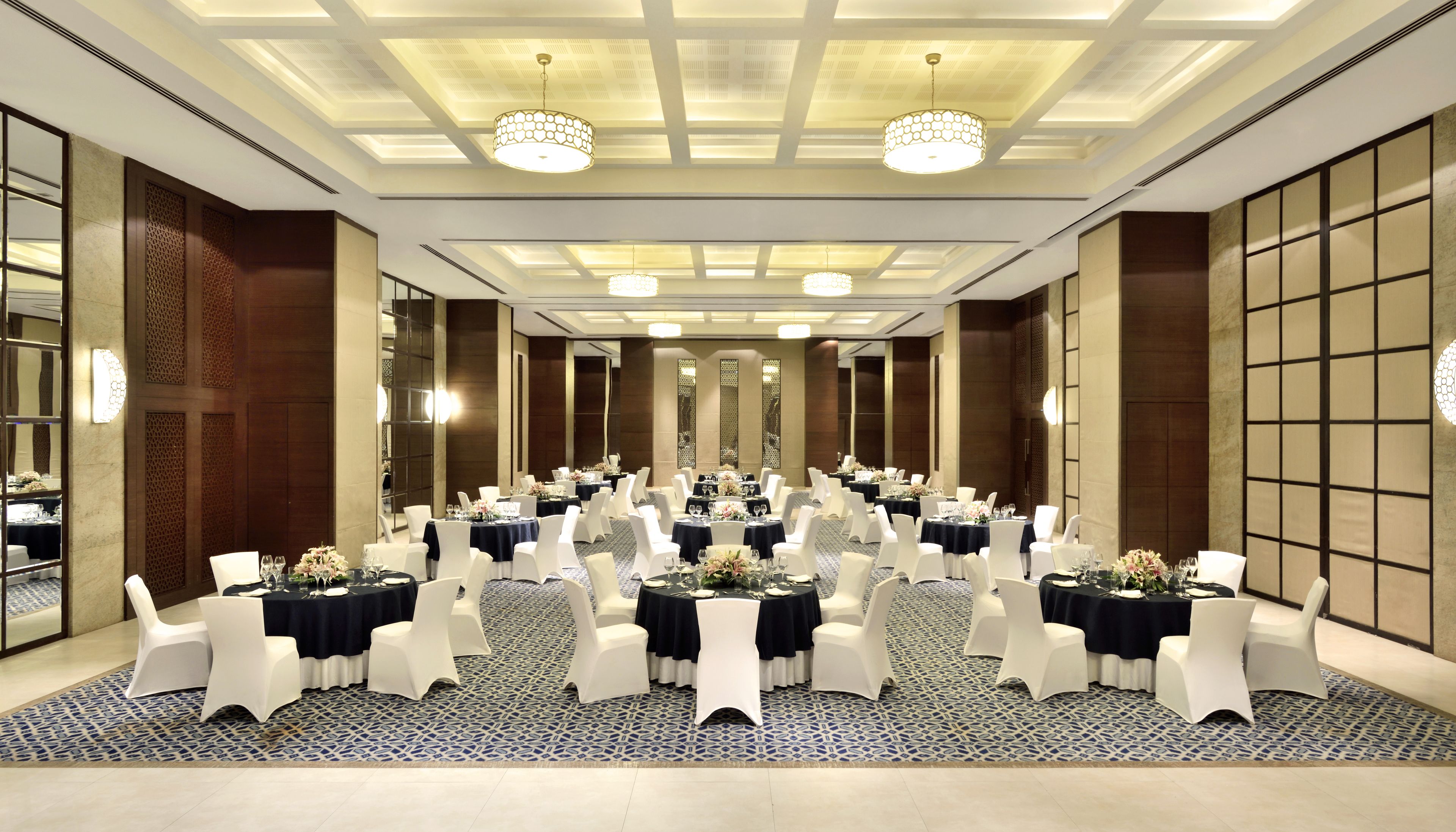 A spacious banquet hall with round tables, white chairs, and a patterned carpet.