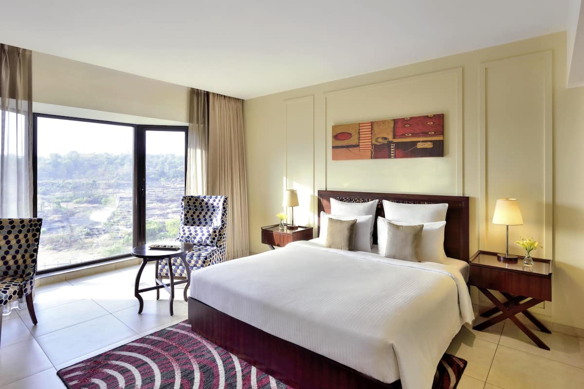 Una habitación de hotel con una cama grande, vista a un paisaje, una silla de lunares y una mesita de noche con una lámpara.