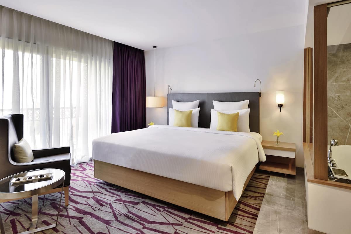 Habitación de hotel con una cama grande, una mesita de noche, una silla, una lámpara y un baño con espejo y lavamanos.