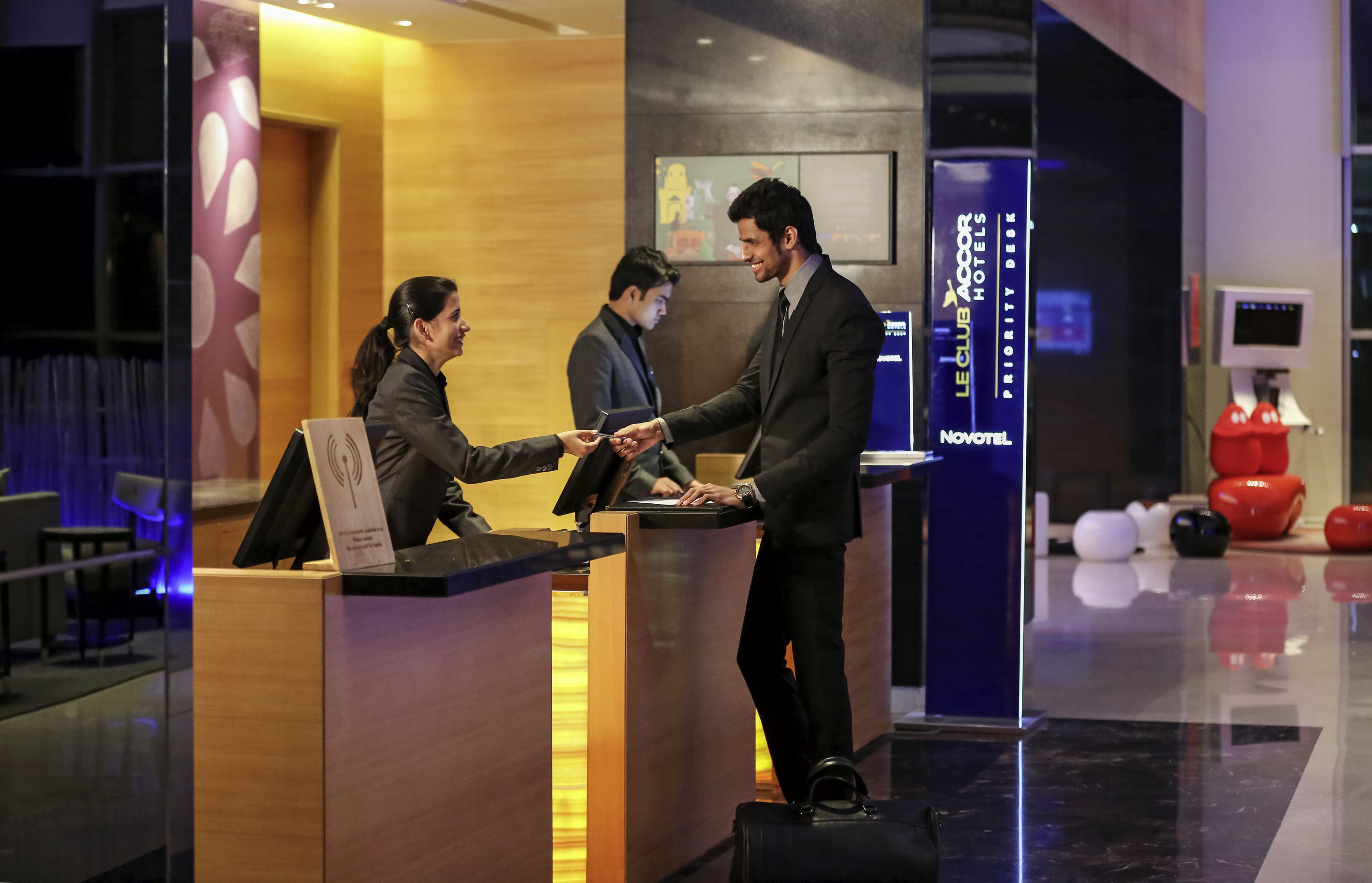 A hotel reception area with a wooden reception desk, a digital screen, and a Novotel logo.