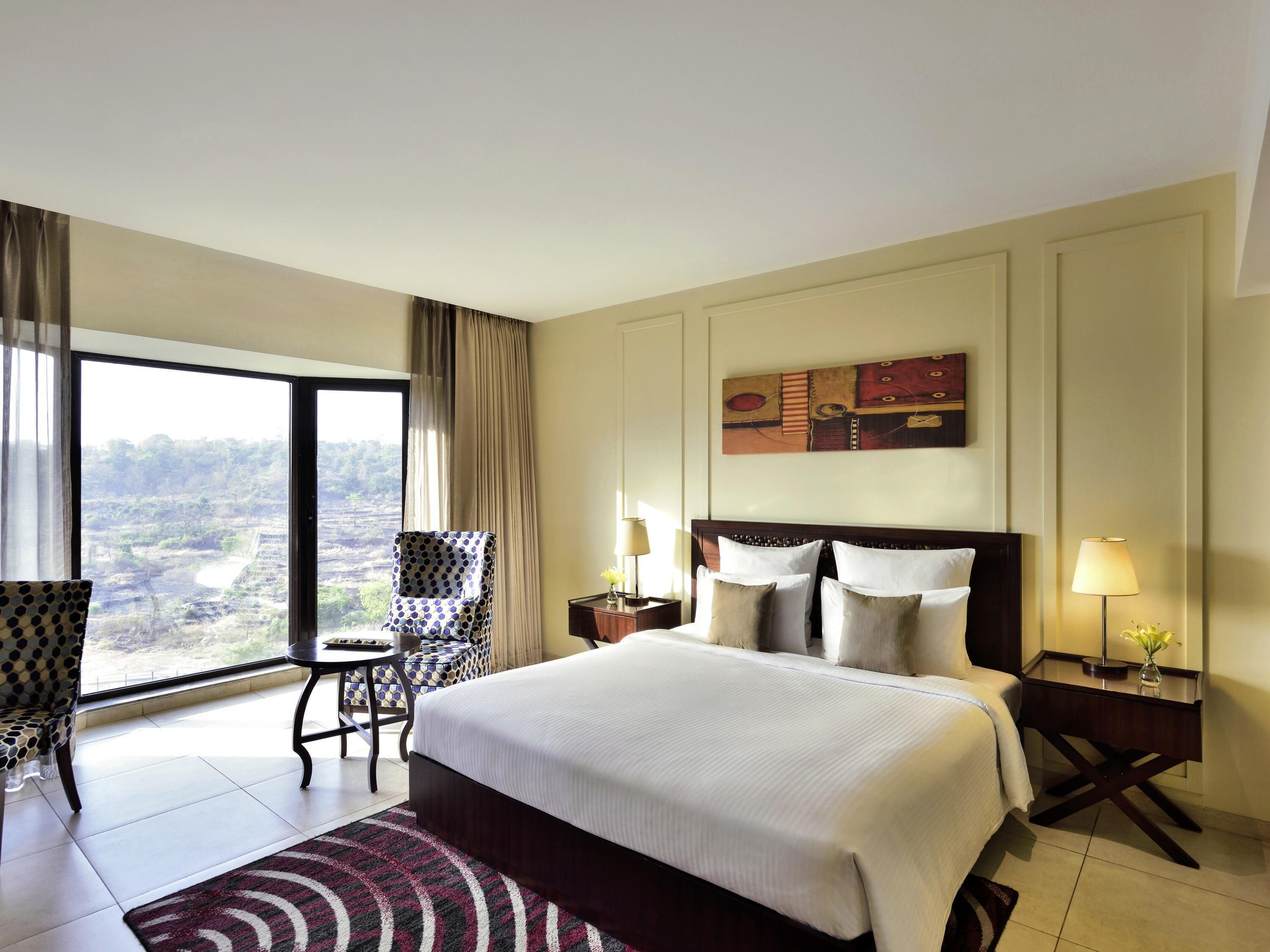 A hotel room with a large bed, a chair, a small table, a nightstand, and a view of a landscape.