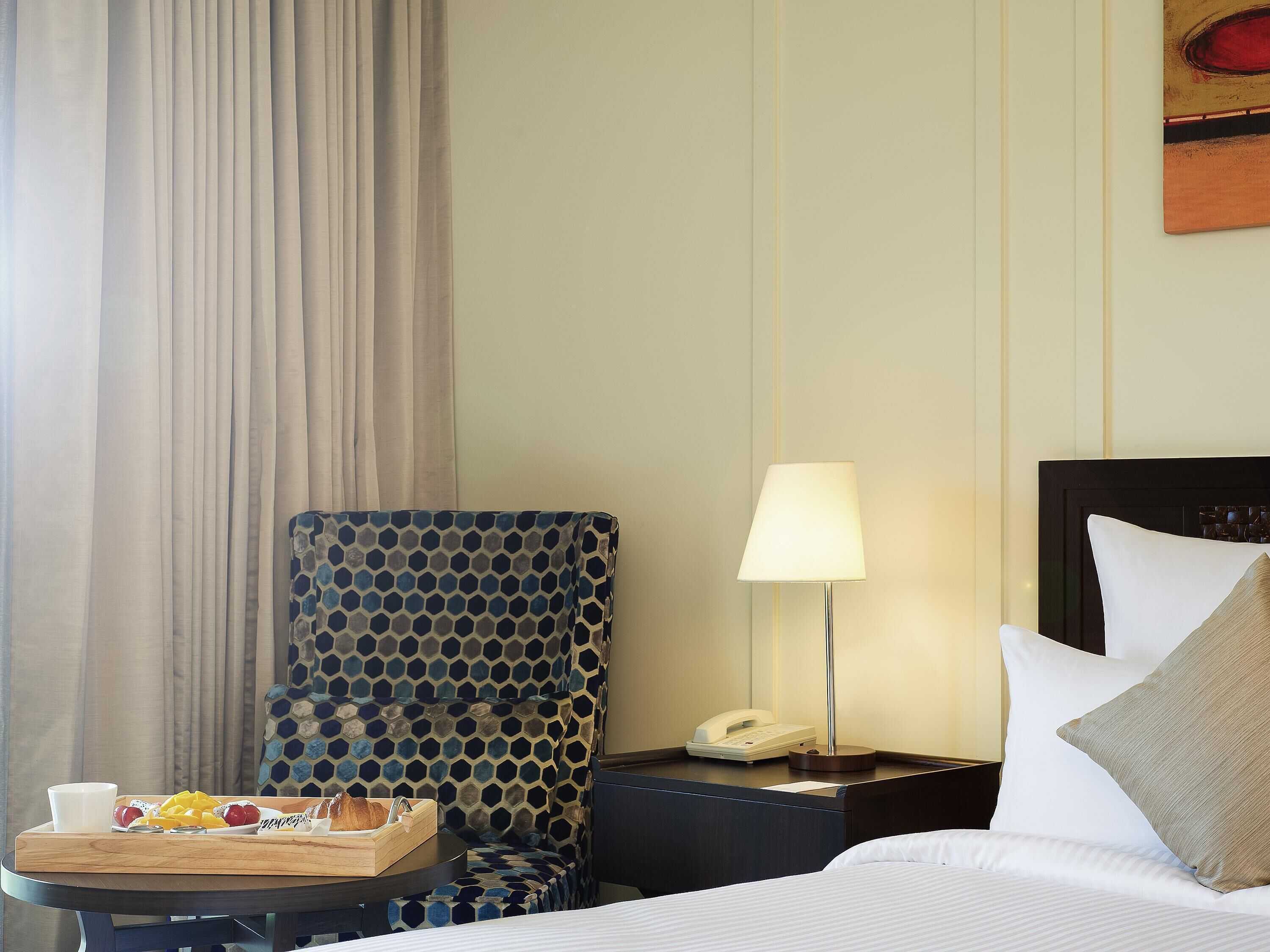 A hotel room with a bed, a bedside table with a lamp, a chair, and a tray with breakfast items.