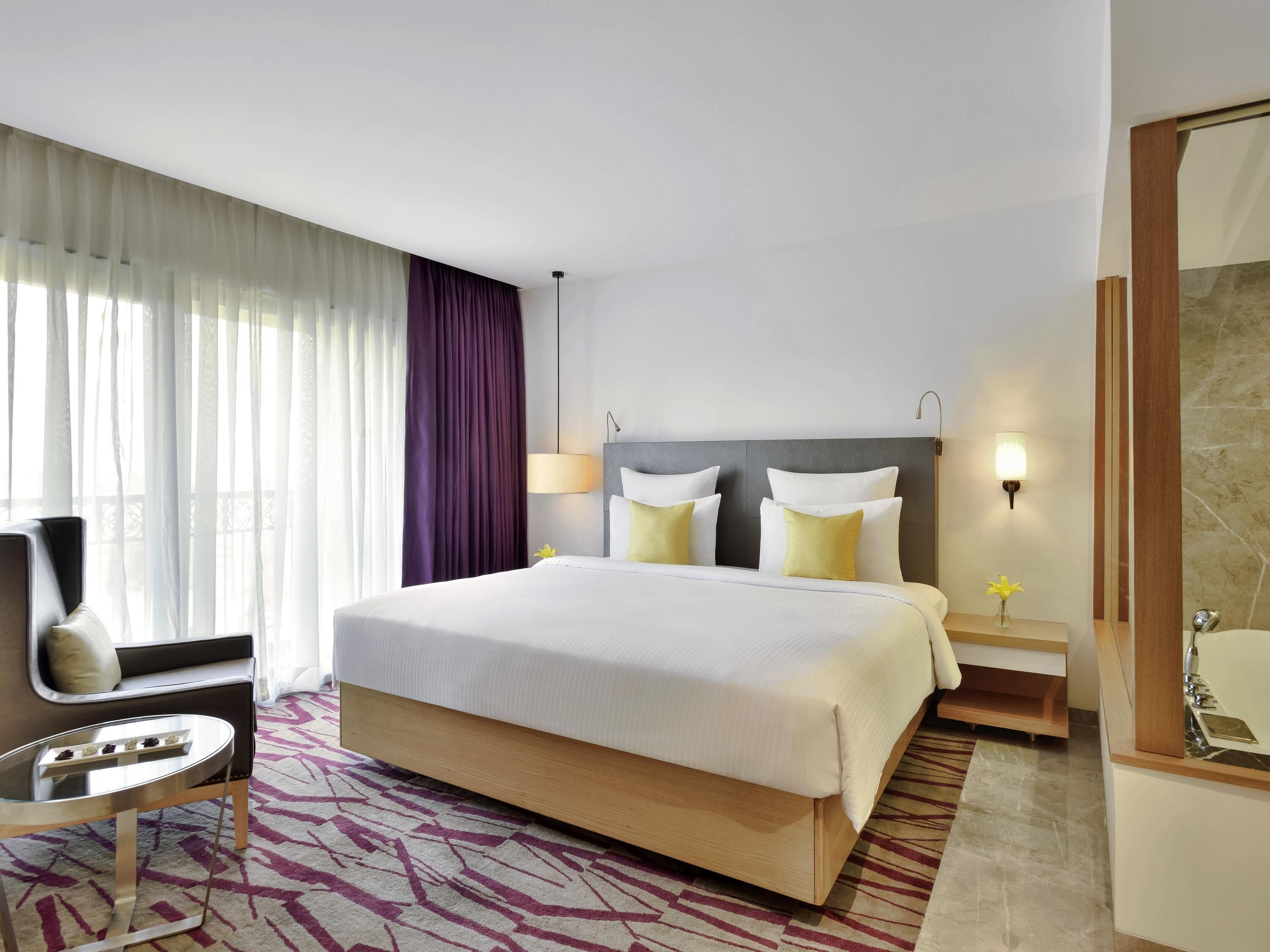A hotel room with a large bed, bedside tables, a chair, a bathroom with a mirror and sink, and a window with curtains.