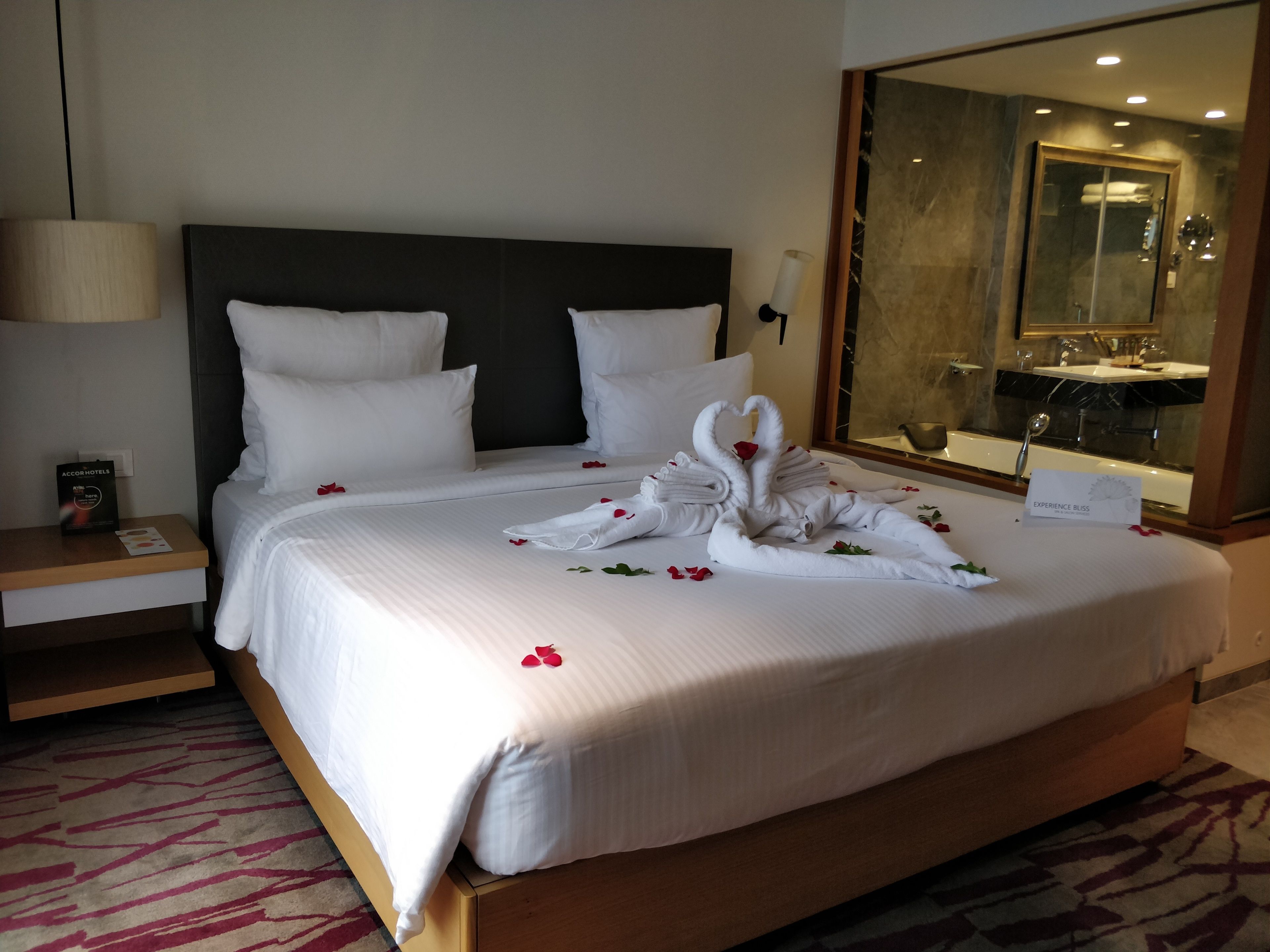 A hotel room with a neatly made bed, a nightstand with a lamp, and a large mirror reflecting a bathroom.