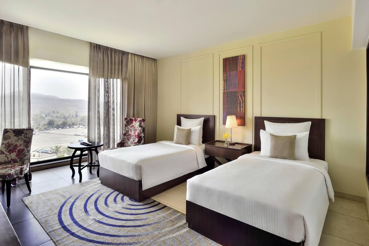A hotel room with two beds, a desk, a chair, a view of a parking lot, and a large window with curtains.