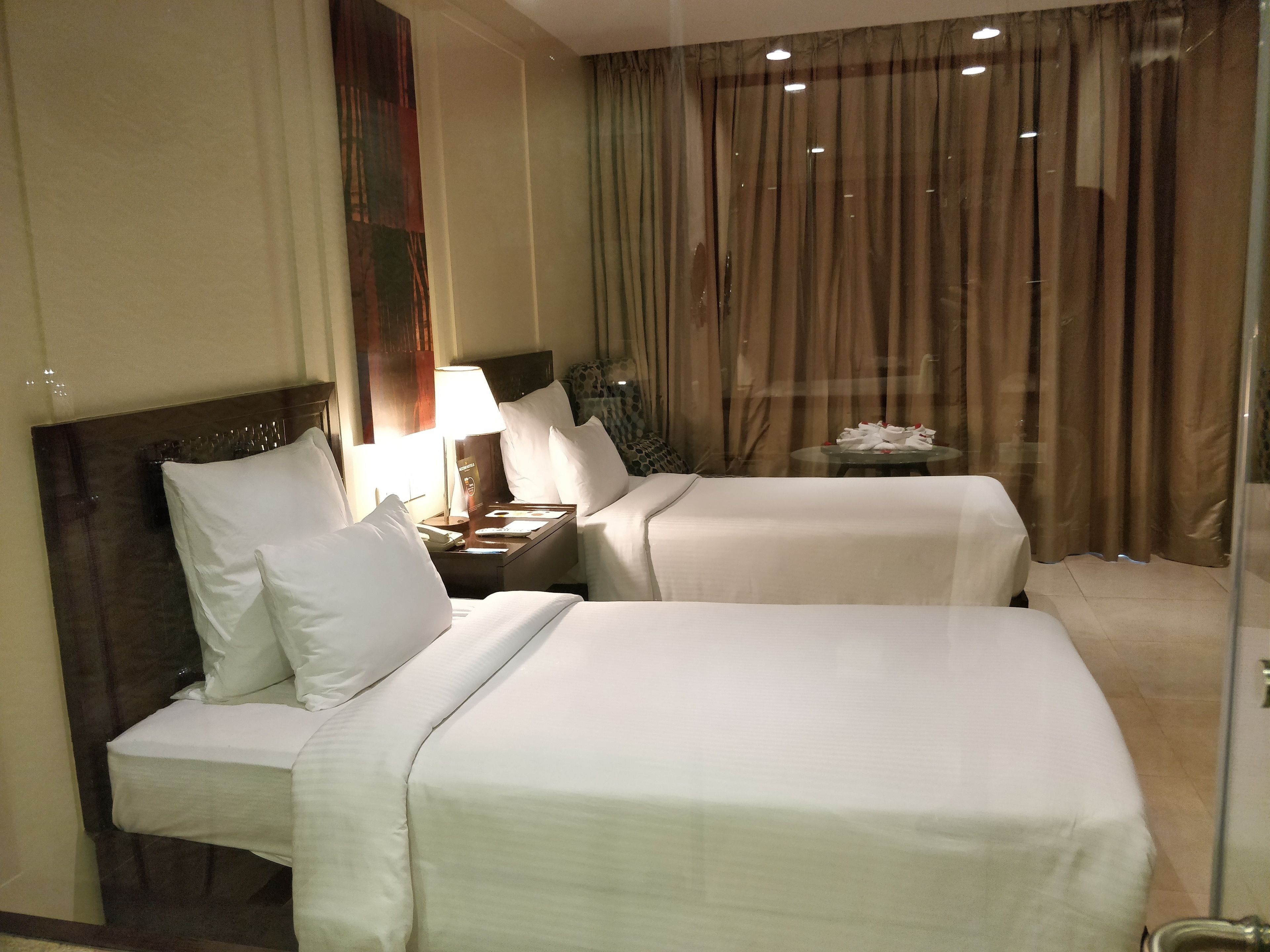 A hotel room with two beds, a nightstand with a lamp, and a view of the city through curtains.