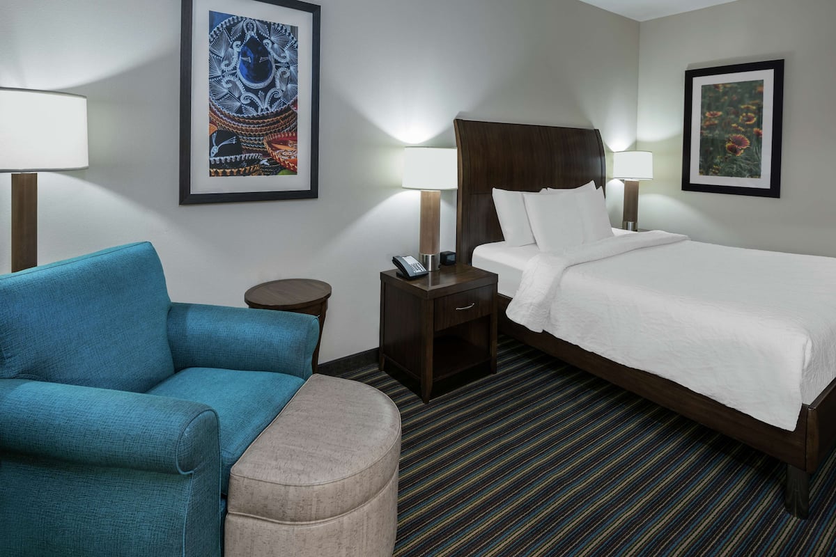 A hotel room with a bed, a blue armchair, a small round table, and two wall-mounted lamps.