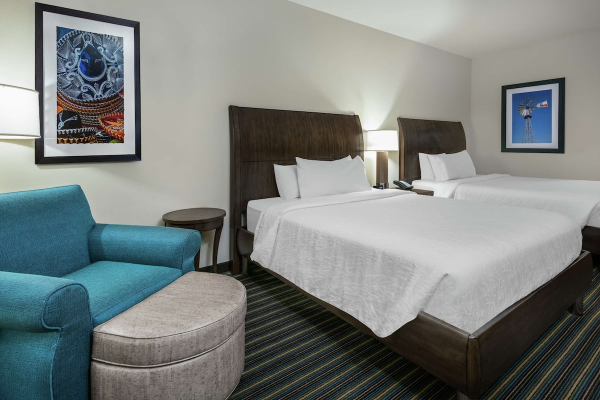 A hotel room with two beds, a blue armchair, a round ottoman, and two framed pictures on the walls.