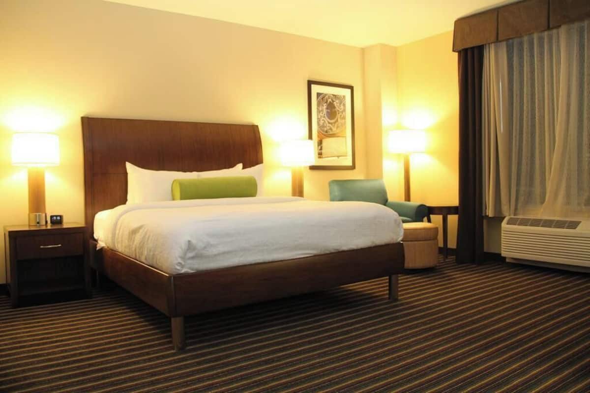 A hotel room with a large bed, two bedside tables with lamps, a chair, a small table, and a window with curtains.