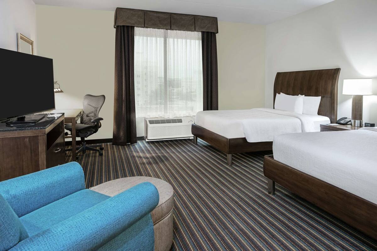 A hotel room with a bed, a desk with a TV, a chair, and a window with curtains.