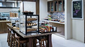 A modern kitchen with a central island, bar stools, a chalkboard with a message, and various food items. - Hilton Garden Inn San Antonio At The Rim (San Antonio)