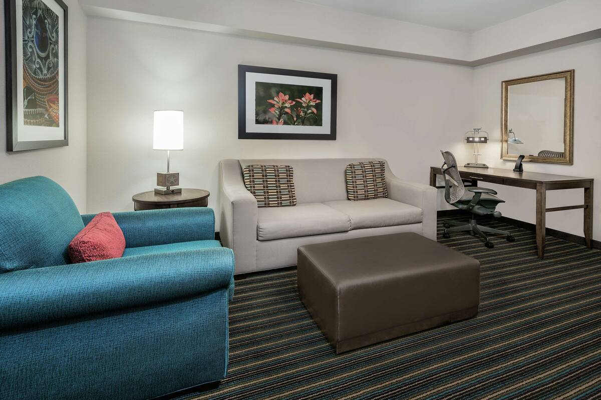 A hotel room with a sofa, a chair, a desk with a chair, a lamp, a framed picture, and a large mirror.