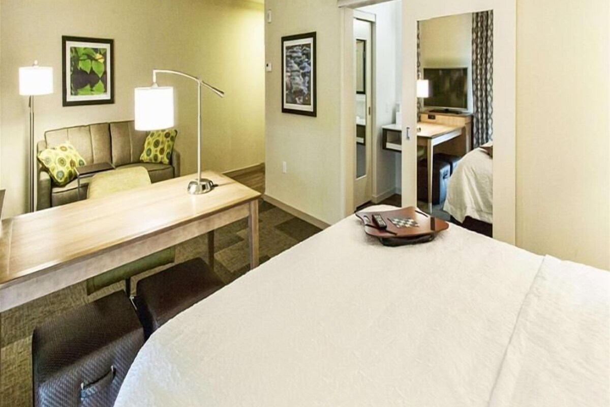 A hotel room with a bed, a desk, a chair, a lamp, and two framed pictures on the wall.