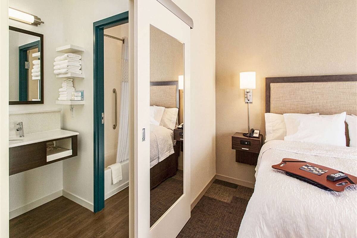 A hotel room with a bed, bedside table, lamp, and a bathroom with a sink and mirror.
