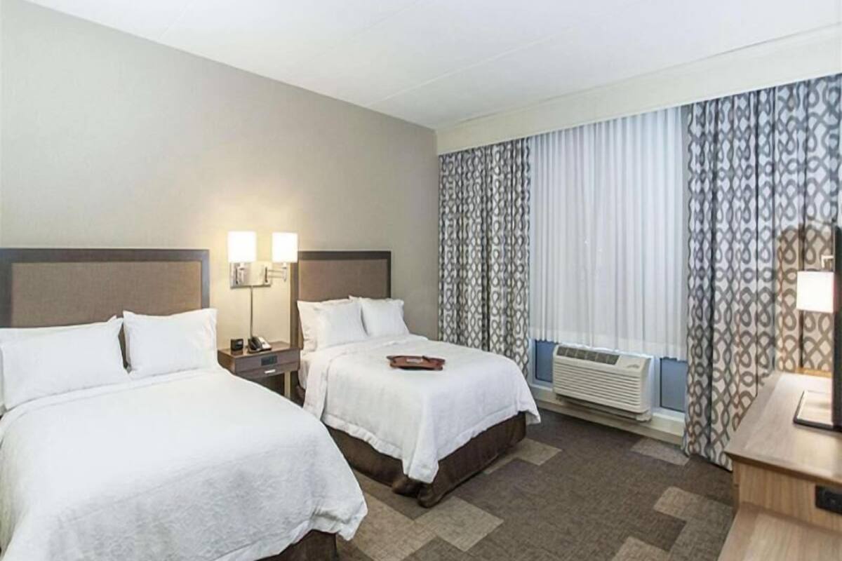 A hotel room with two beds, a bedside table with a lamp, a window with curtains, and an air conditioning unit.