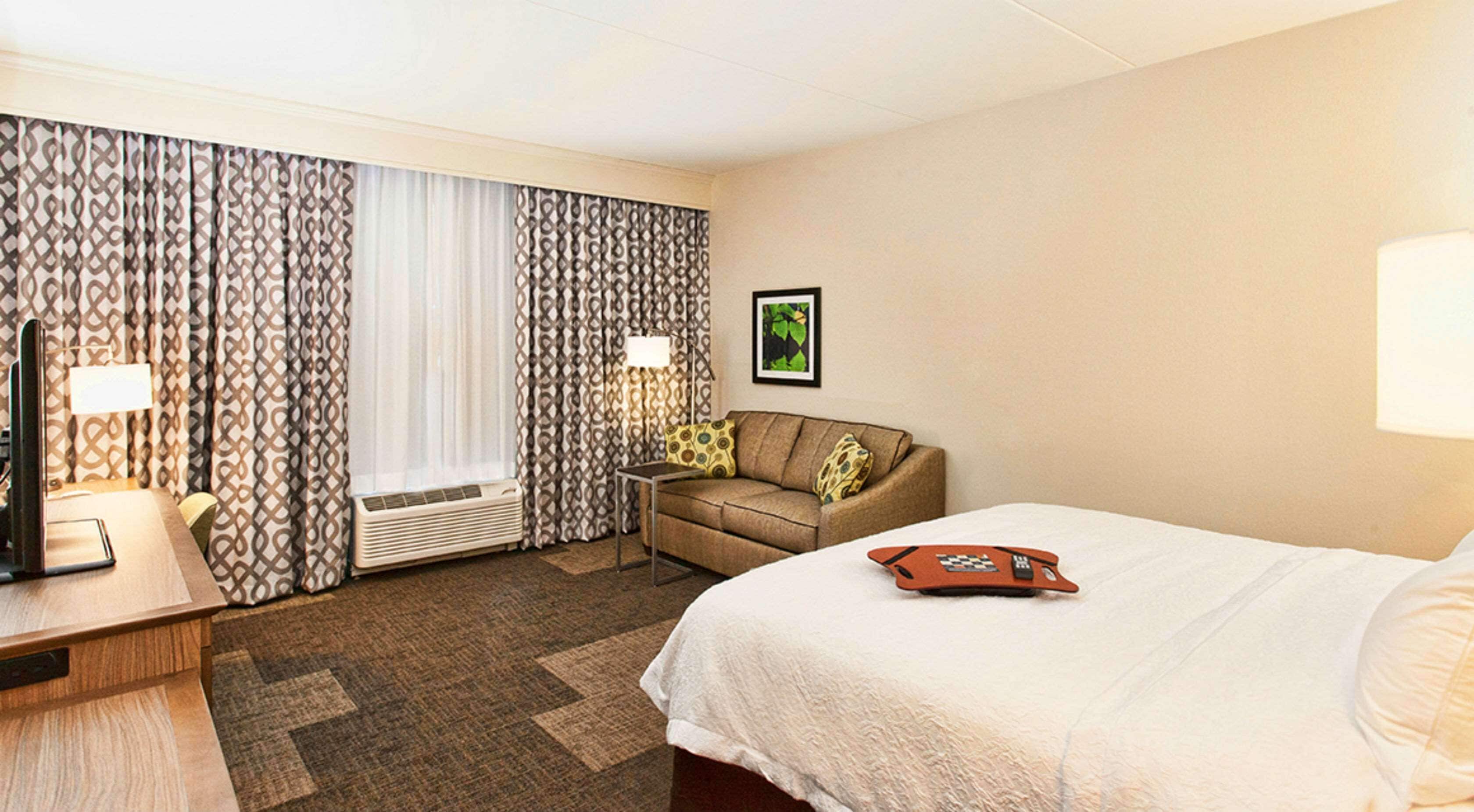 A hotel room with a bed, a desk with a lamp, a sofa, and a window with patterned curtains.