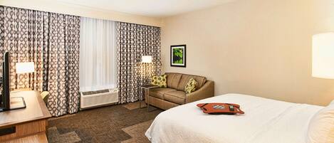 A hotel room with a bed, a desk with a lamp, a sofa, and a window with patterned curtains.