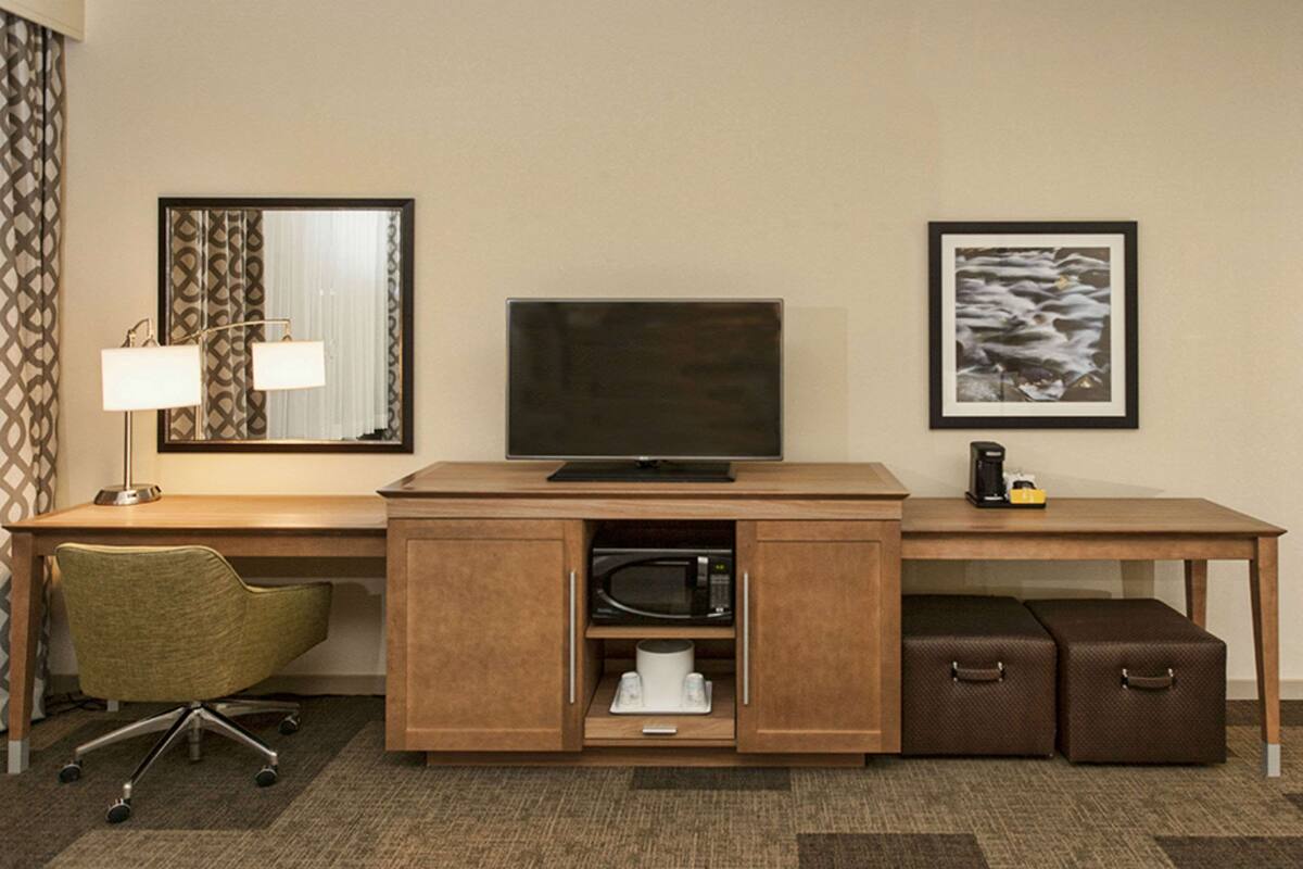 A hotel room with a wooden desk, a television, a microwave, a coffee maker, a green office chair, and two brown suitcases.