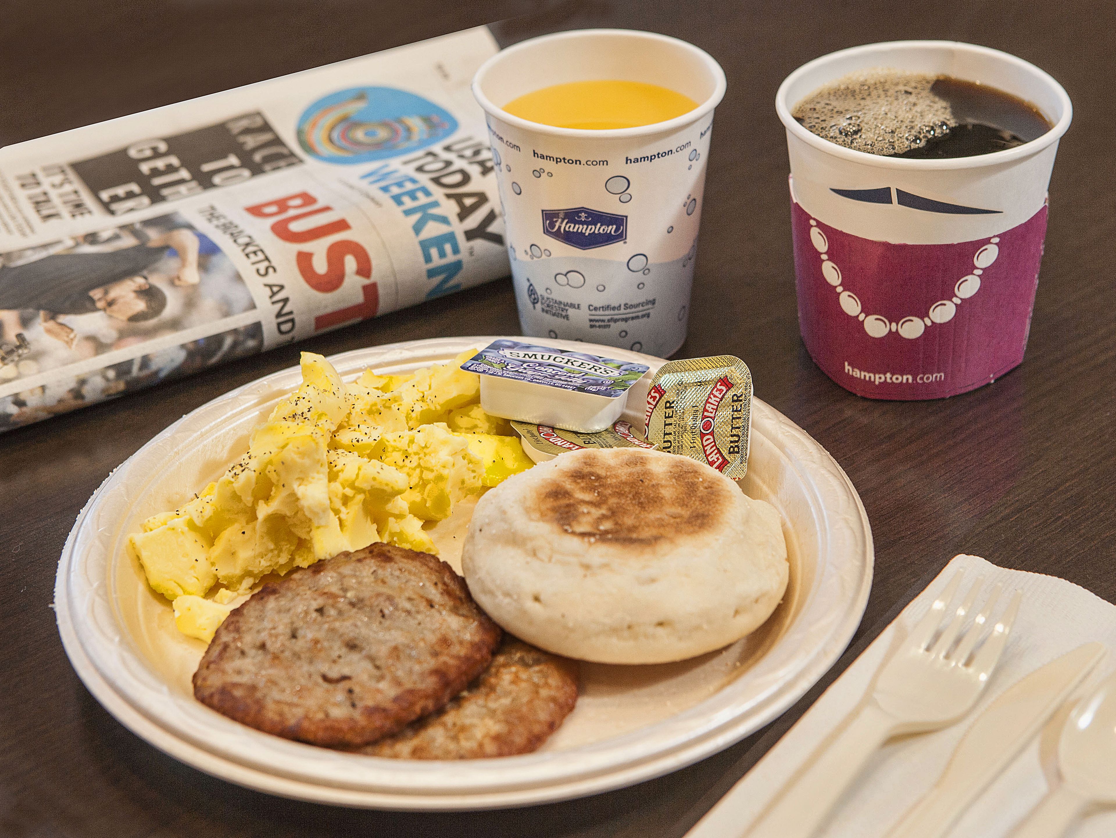 A breakfast plate with scrambled eggs, sausage, and a biscuit, accompanied by a cup of coffee and a newspaper.
