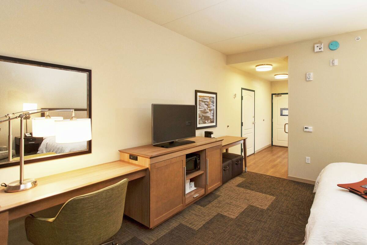 A hotel room with a bed, a desk with a computer, a chair, a TV, and a mirror.