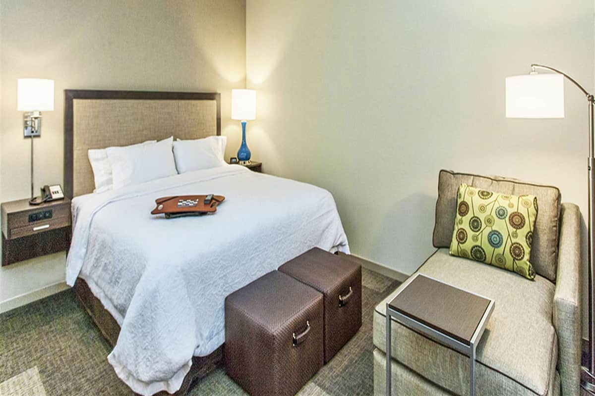 A hotel room with a bed, a nightstand, a lamp, a small table, a sofa, and a suitcase.