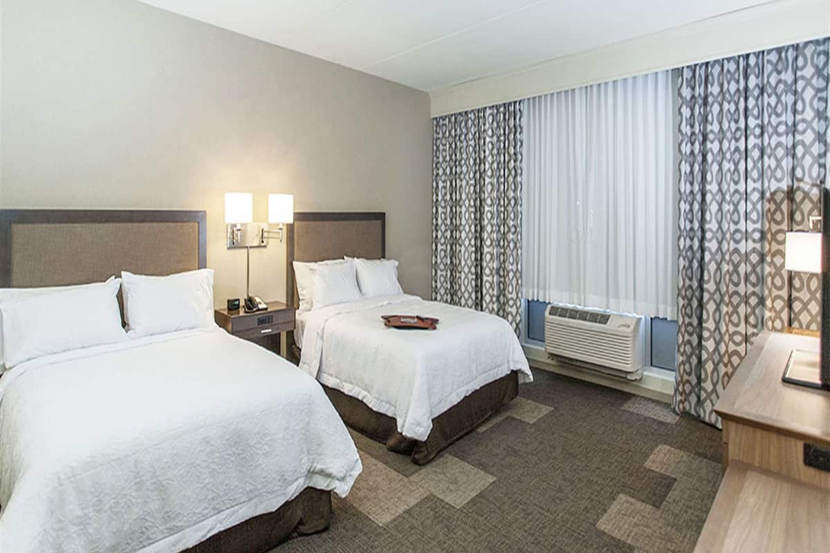A hotel room with two beds, a bedside table with a lamp, a window with curtains, and a wall-mounted air conditioner.