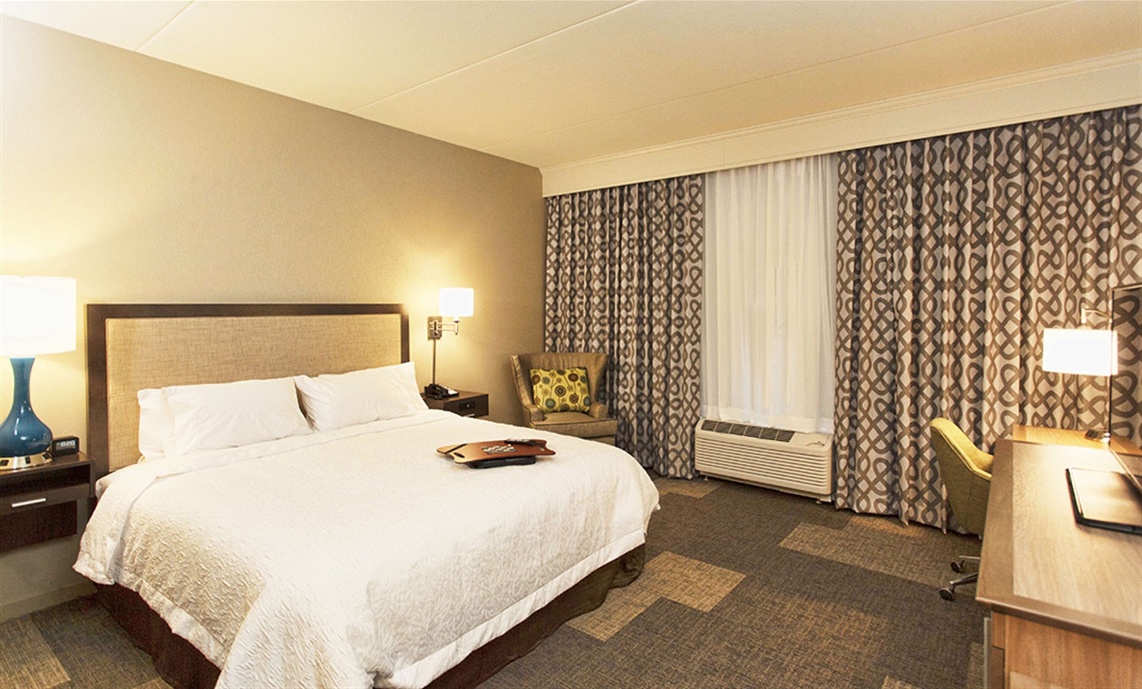 A hotel room with a large bed, a desk, a chair, a lamp, and a window with patterned curtains.