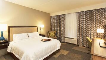 A hotel room with a large bed, a desk, a chair, a lamp, and a window with patterned curtains.