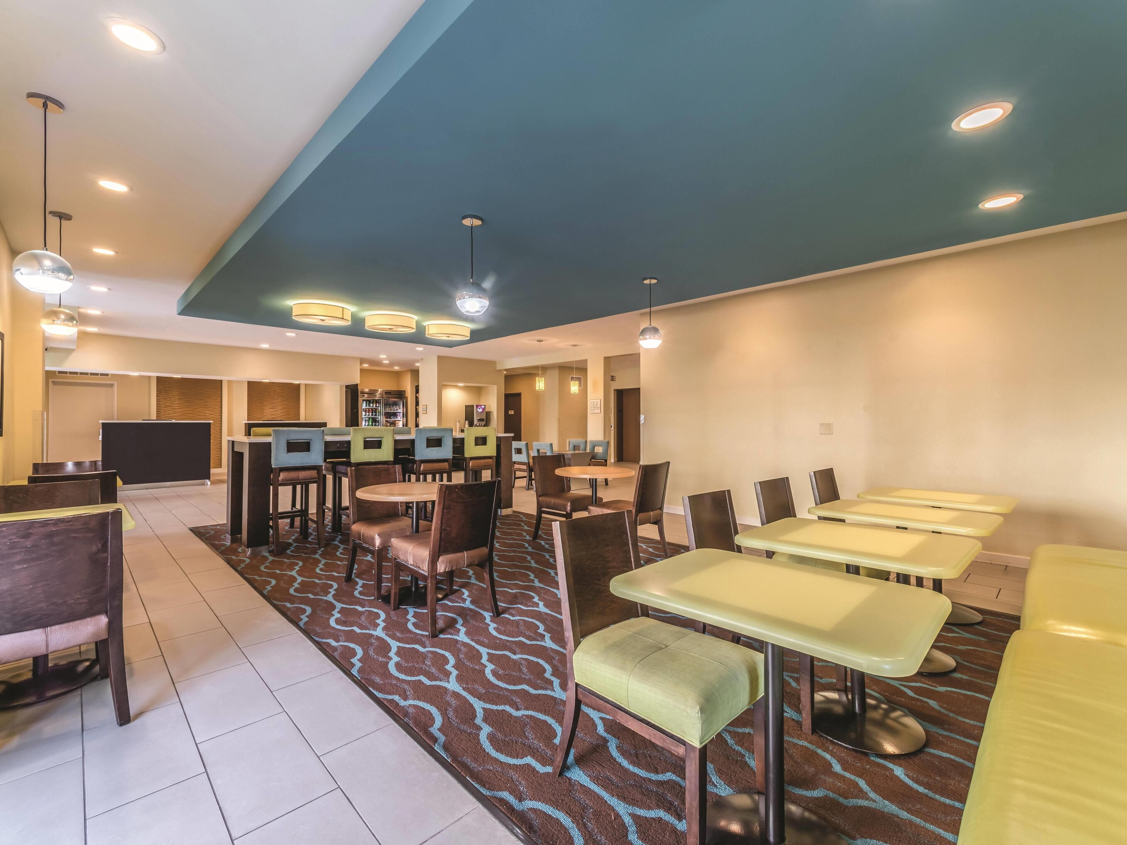 A hotel dining area with multiple tables and chairs, a reception desk, and a kitchen in the background.