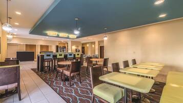 A hotel dining area with multiple tables and chairs, a reception desk, and a kitchen in the background.