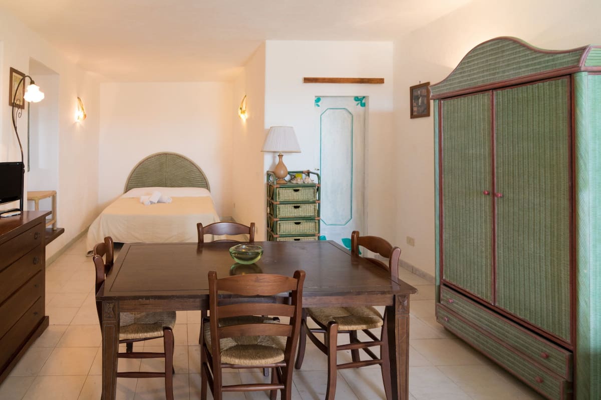 Une pièce comprenant un lit, une table à manger en bois avec des chaises, une lampe, une armoire et un téléviseur posé sur une commode.