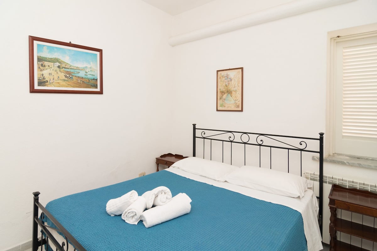 Une chambre à coucher comprenant un lit, une table de chevet, une fenêtre avec des stores et deux tableaux encadrés accrochés au mur.