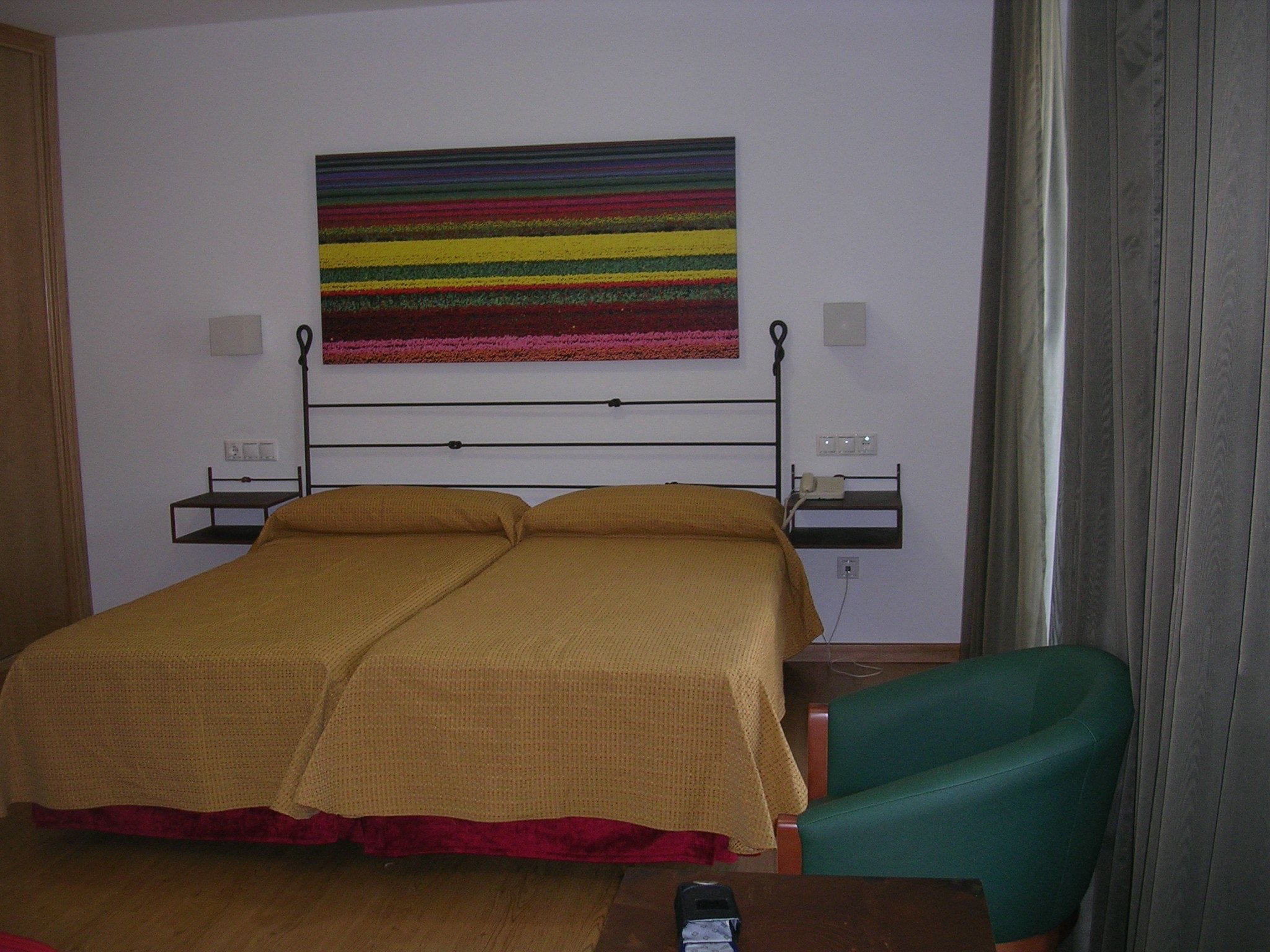 A hotel room with a bed, a green chair, a bedside table, and a colorful striped artwork on the wall.