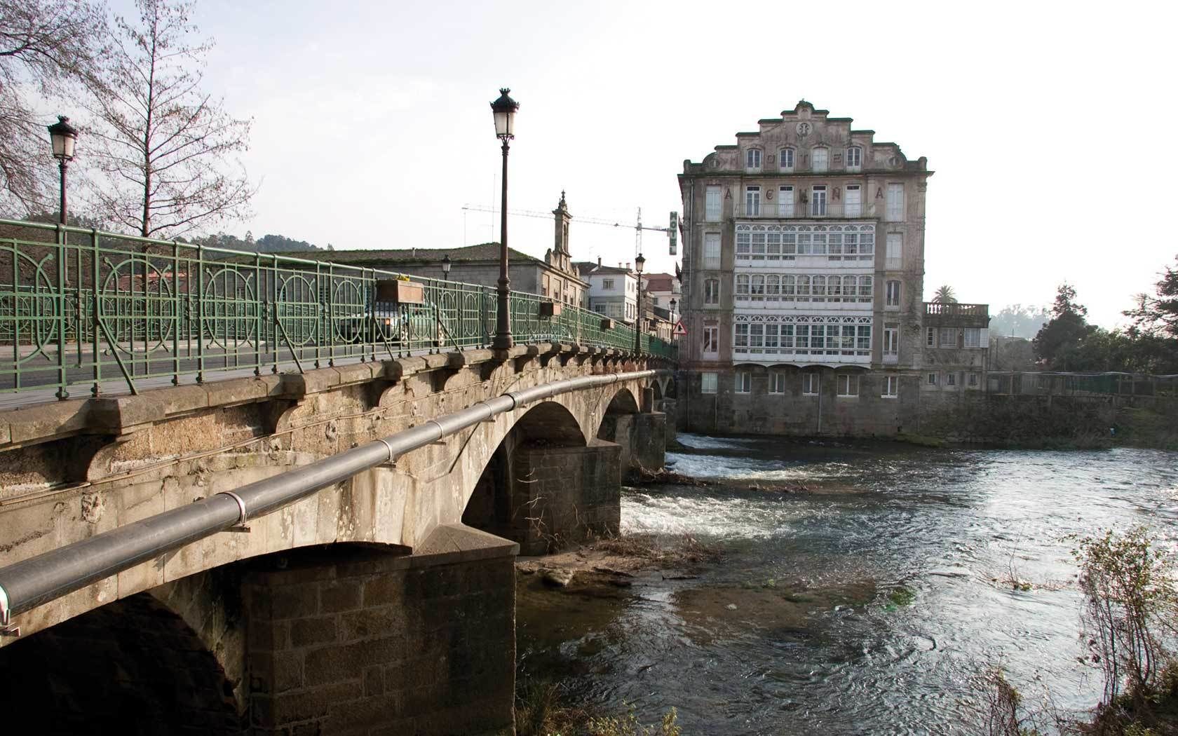 A stone bridge with a metal railing over a river, leading to a historic building with multiple windows and a clock tower.